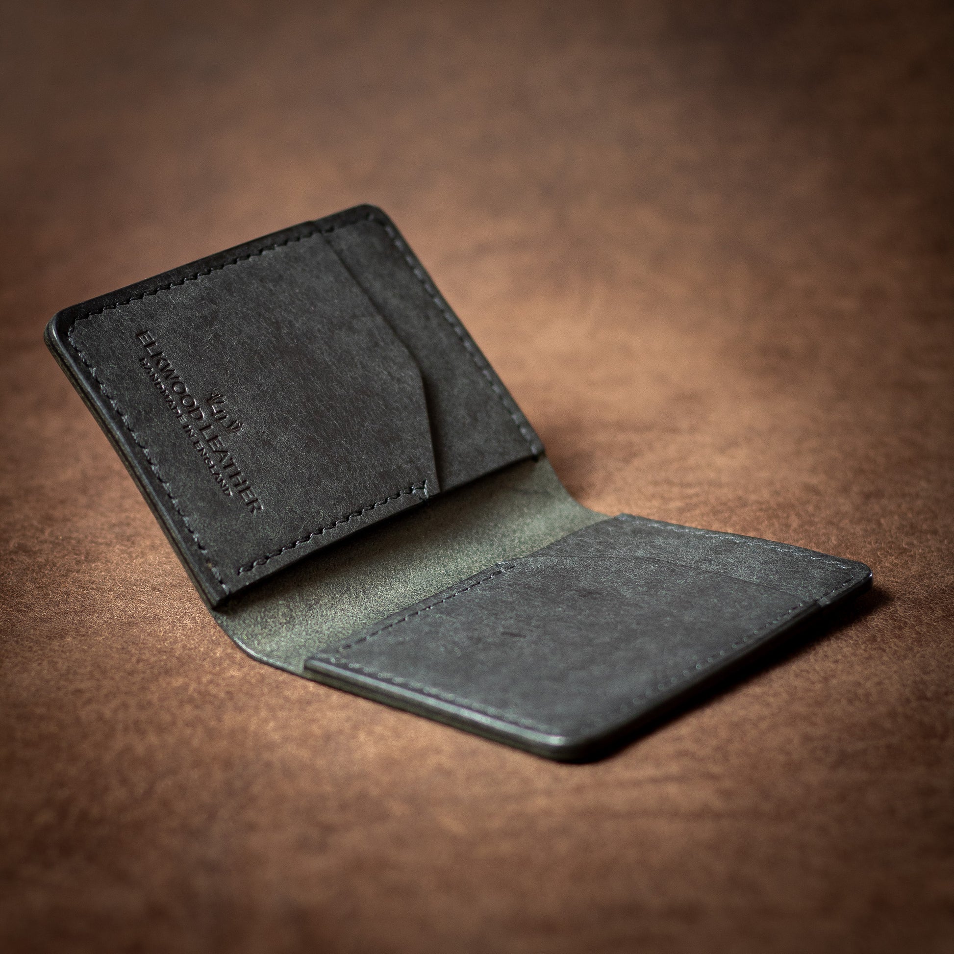 Elkwood Leather black leather bifold cardholder wallet with black stitching on a brown leather background open